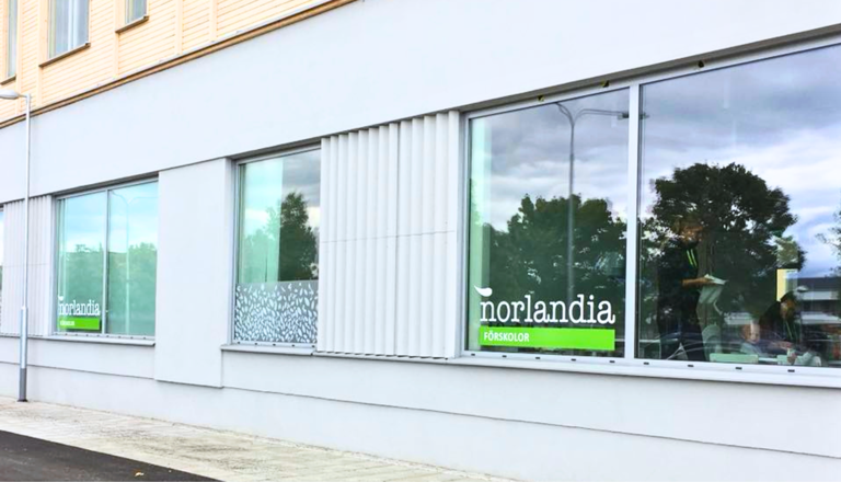 Förskolebyggnad i ljus fasad med stora fönster och Norlandia Förskolor-logga tydligt synlig från gatan.