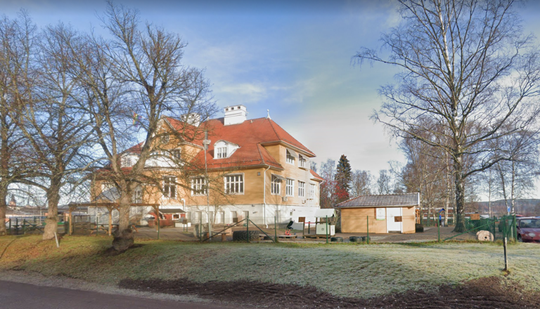 Charmig förskolebyggnad i klassisk stil, omgiven av stora träd och lekyta – en plats för trygghet, glädje och upptäckter.