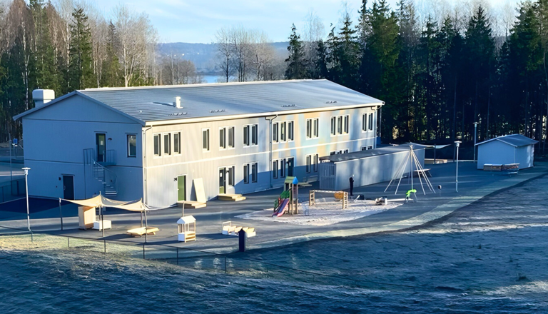 Ljus och modern förskolebyggnad i grått, omgiven av natur – en trygg plats för lek, utforskande, glädje och nya upptäckter.