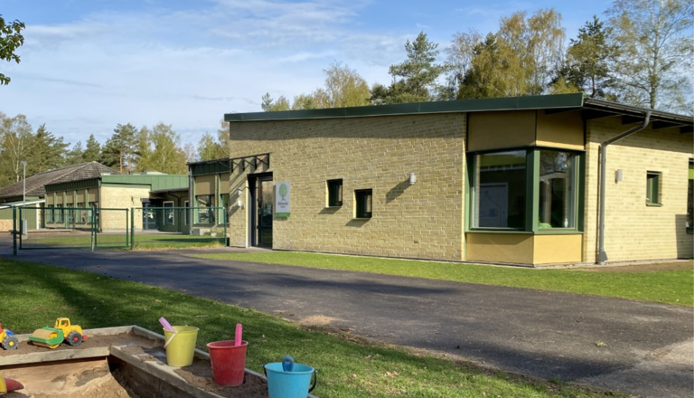 Ljusgul tegelbyggnad med gröna detaljer, grönskande omgivning och sandlåda med leksaker i förgrunden.