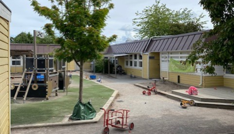 Gul enplansförskola med brunt tak, lekplats med klätterställning, cyklar och trädgård i förgrunden.