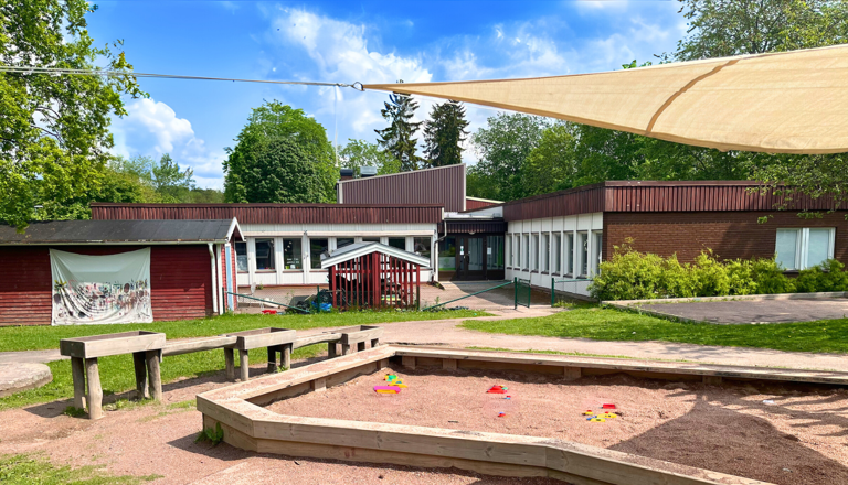 Förskolegård med sandlåda, lekredskap och grönska framför låg brun- och vit byggnad – plats för lek och trygghet.