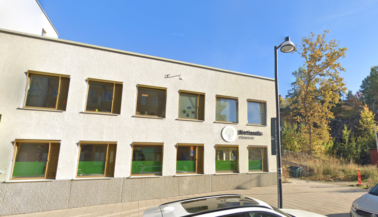Tvåvåningsförskola med ljus fasad och stora fönster i stadsmiljö – en trygg plats för gemenskap, lek, lärande och nyfikenhet.