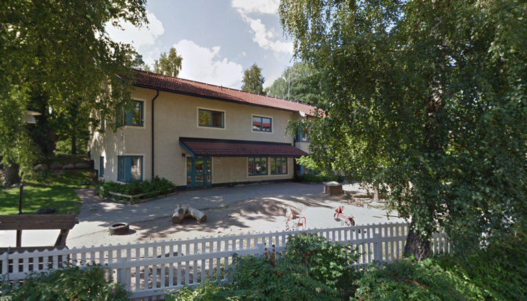 Förskolebyggnad i två våningar med lekcyklar och träd på gården – en trygg, grön plats för lek, gemenskap och upptäckarglädje