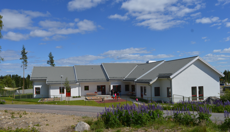 Vit förskolebyggnad med grått tak, barn som leker på gården och blå himmel i bakgrunden.