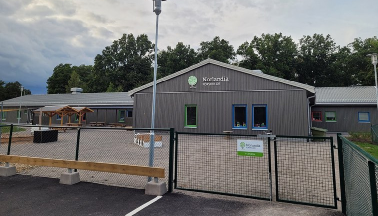 Modern förskola med grå fasad och färgglada fönster, gård med lekyta och skog i bakgrunden – en plats för lek och lärande.