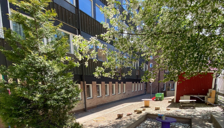 Grönskande innergård med sandlåda, stubbar och lekytor intill förskolefasad – en trygg plats för utforskande lek.