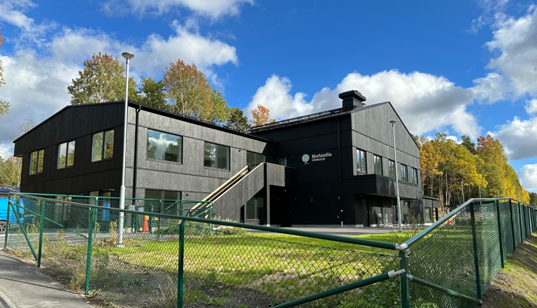 Svart tvåvånings förskola med Norlandia-skylt, inhägnad gård och skog intill – en trygg plats för lek, lärande och gemenskap.