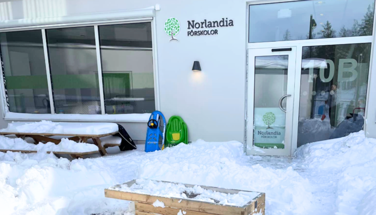 Entré till förskolebyggnad med Norlandia-logotyp, snötäckt mark, pulkor och bord utanför glasdörren.