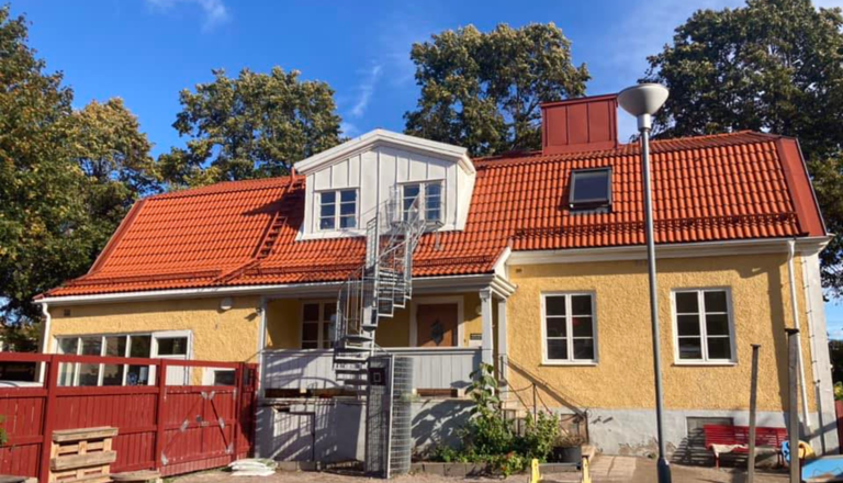 Gul tvåvånings förskolebyggnad med rött tegeltak och spiraltrappa, omgiven av träd och blå himmel.
