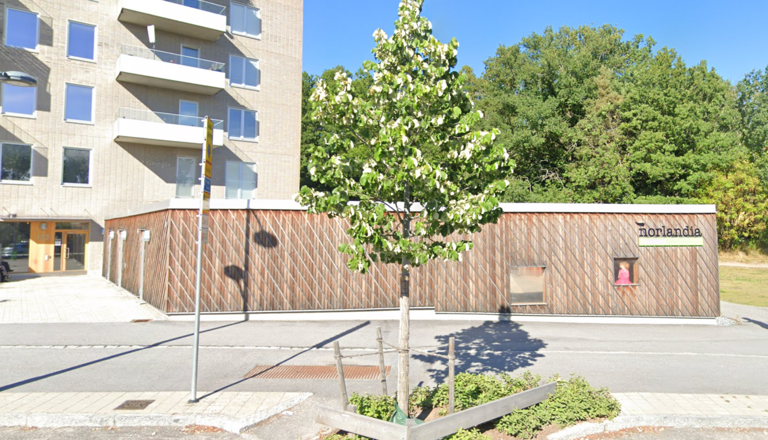 Träklädd förskolebyggnad med Norlandia-logotyp, grönskande träd och bostadshus – en trygg plats i stadens närhet.
