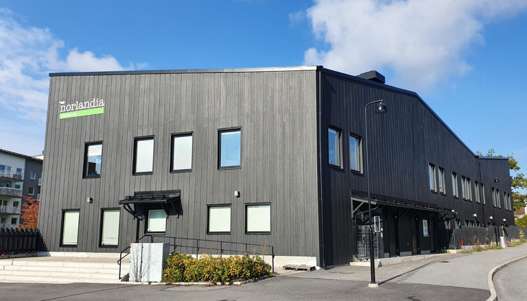 Tvåvånings förskolebyggnad i mörk träfasad med Norlandia Förskolor-logga och flera entréer.