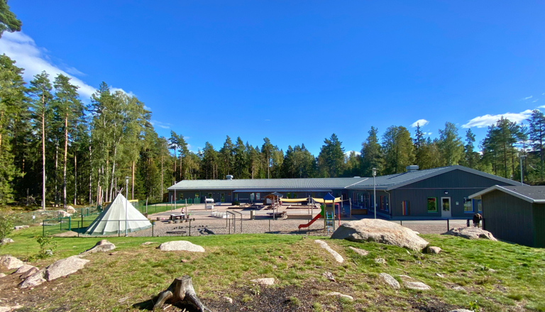 Låg enplansförskola med grå träfasad och färgglada fönsterkarmar, omgiven av skog och med asfalterad entrégård.