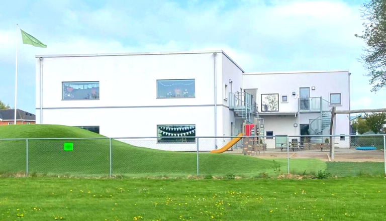 Modern tvåvånings förskolebyggnad i vitt med lekplats, spiraltrappa och en grön flagga som vajar vid entrén.