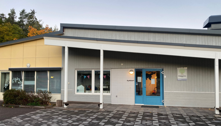 Grå och gul byggnad med blå dörrar och skylt för Norlandia Förskolor Skogsgläntan, dekorerad med vimplar i fönstret.