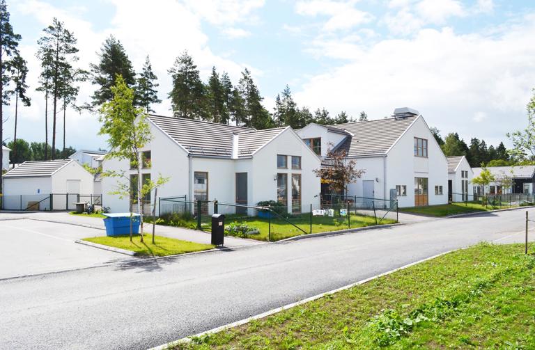 Modern förskolebyggnad i vit puts med höga fönster, lekytor och naturnära läge omgiven av skog och grönska.