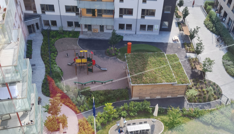 Innergård med förskolebyggnad, grönskande tak, lekplats med rutschkana och sittplatser för utevistelse.