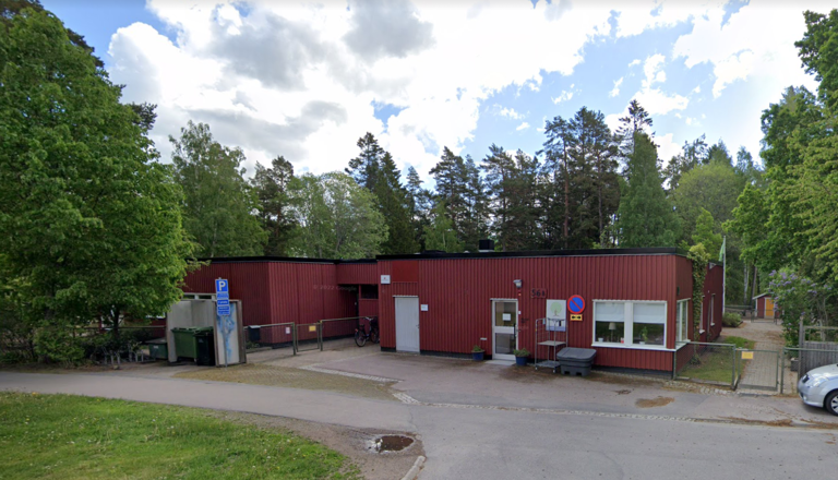 En röd enplansbyggnad med vit dörr och Norlandia Förskolor-skylt, omgiven av grönskande träd och cykelparkering.