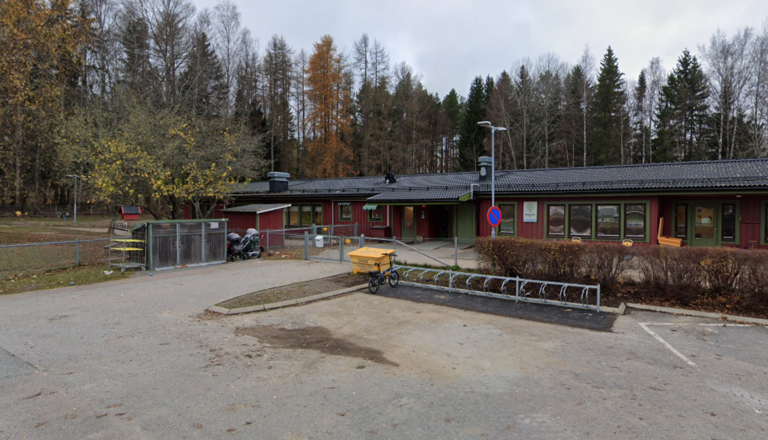 Röd förskolebyggnad vid skogsbryn med cykelställ och höstträd – en trygg, naturnära plats för lek, lärande och gemenskap.