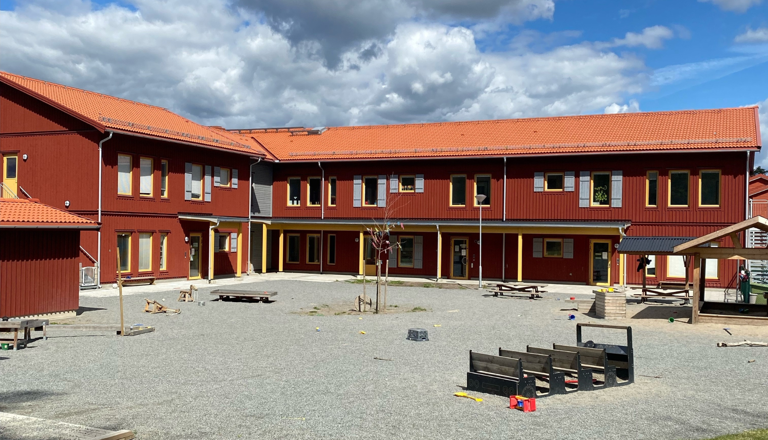 Röd tvåvåningsförskola med orange tak och stor gård fylld med lekutrustning under en blå himmel med moln.