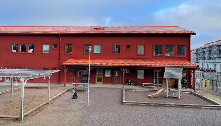 Röd förskolebyggnad i två plan med lekställning, sandlådor och grusgård framför entrén.