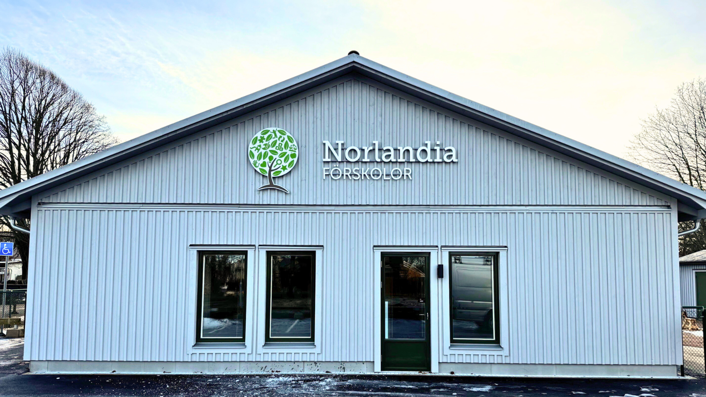 Ljusgrå förskolebyggnad i trä med sadeltak, tre fönster och en dörr på framsidan samt Norlandia Förskolor-logotyp.
