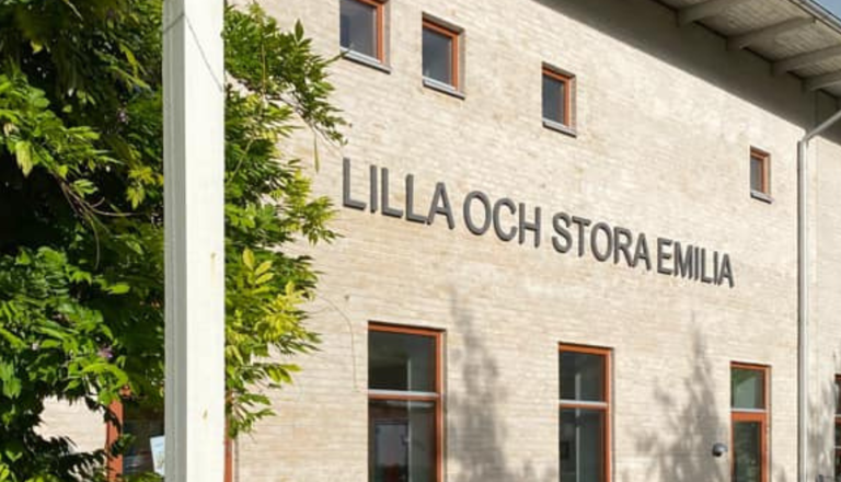 Ljus tegelfasad med stora bokstäver “Lilla och Stora Emilia” och gröna träd – en lugn plats för lek och lärande.