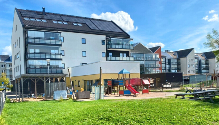 Modern förskola i bottenplan av flerbostadshus med stor gård, lekplats och gräsytor – en trygg plats för rörelseglädje.