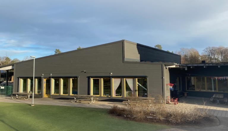Mörk förskolebyggnad i trä med stora fönster och gård med bänkar – en trygg plats för gemenskap, lek och lärande.