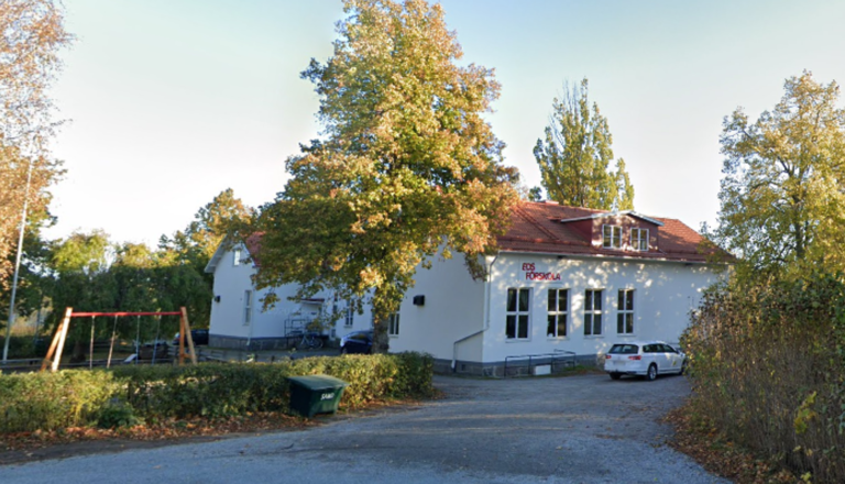 Vit förskolebyggnad med röda takpannor, stora fönster och gungställning – en plats för lek, lärande och gemenskap i grönska.