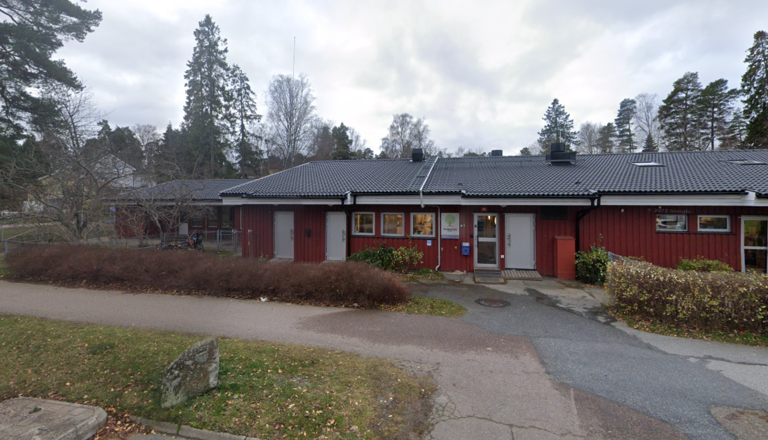 Röd förskolebyggnad i trä med grått tak, omgiven av natur och bostäder – en plats för lek, lärande, nyfikenhet och gemenskap.
