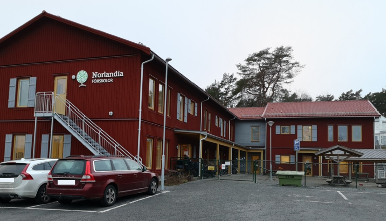 Röd förskolebyggnad i trä med skog i bakgrunden och parkeringsplatser framför – en plats för trygghet, lek och gemenskap.
