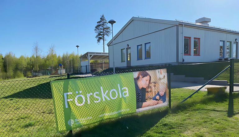 Färgglad förskolebyggnad i gult och grått med staket, barnvagnar och grönska – en trygg plats för lek och lärande.