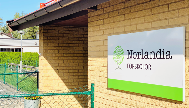 Förskolebyggnad i gult tegel med Norlandia Förskolor-skylt vid entrén och grön nätgrind i förgrunden.