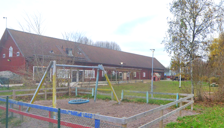 Röd förskolebyggnad i lantlig stil med stor gård, gungställning och lekytor, omgiven av träd och grönska i höstskrud.