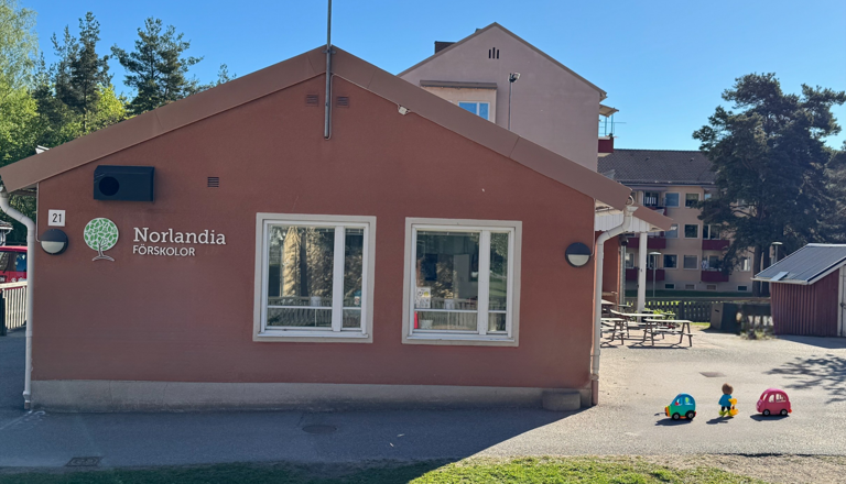 Förskolebyggnad med logotyp, innergård och barn med leksaksbilar – en trygg plats för lek, gemenskap och nyfikenhet.